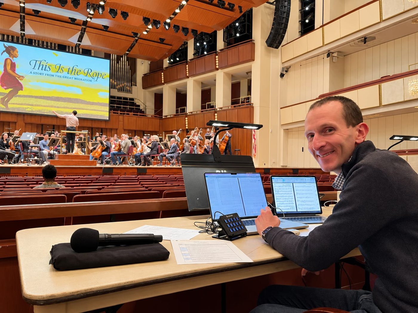 A working desk at the back of a concert hall, with laptops displaying music, a Stream Deck keypad and printed scores, while an orchestra rehearses on stage.